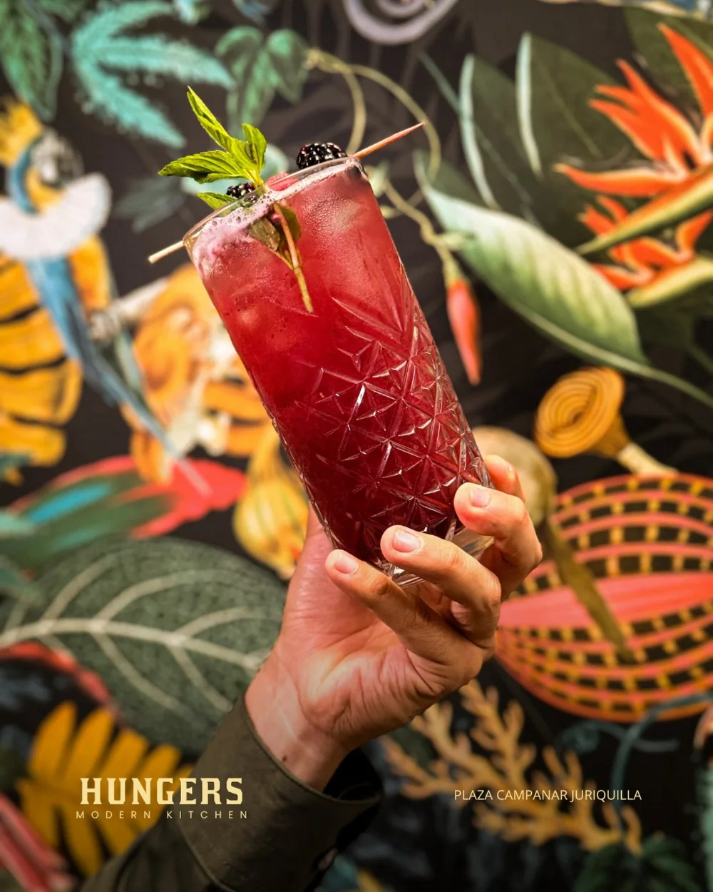Un cóctel rojo en vaso alto tallado, levantado contra el papel tapiz tropical del salón de Hungers.