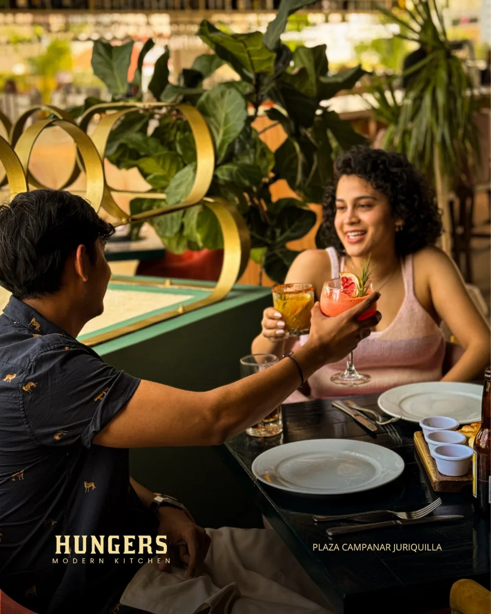 Dos personas brindando con cócteles de la casa en una mesa de Hungers, entre plantas y aros dorados.