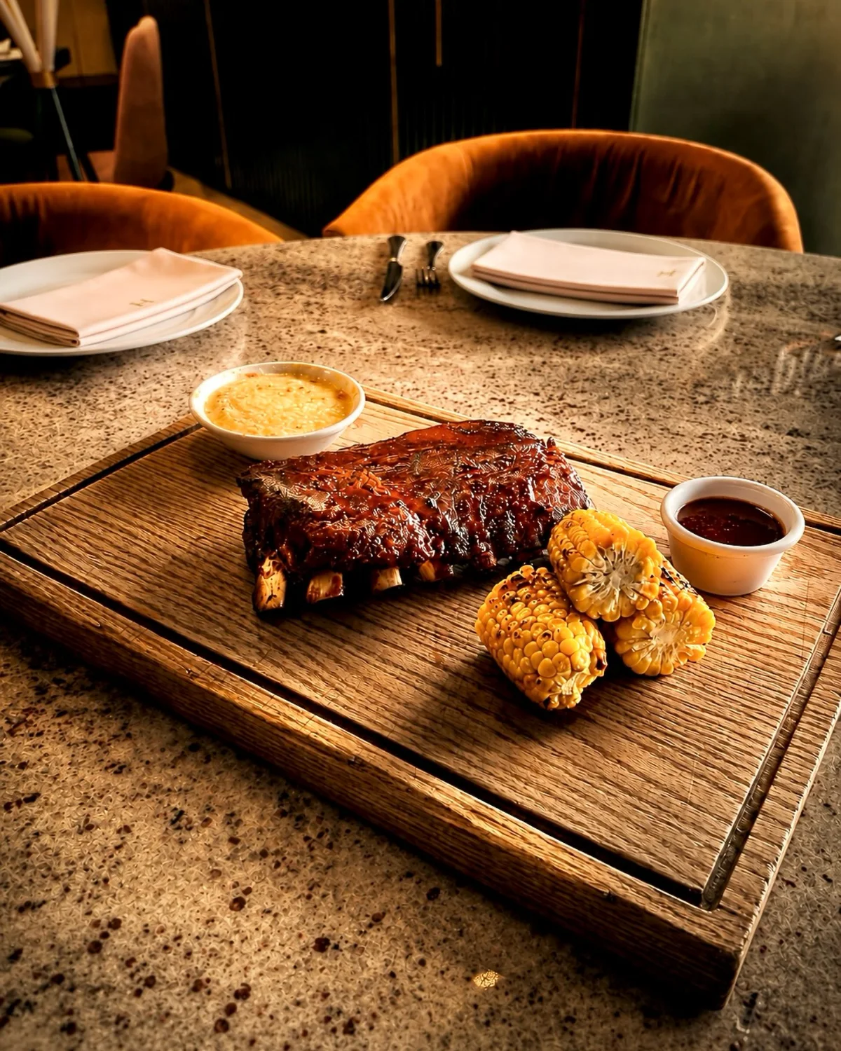 Costillar baby back con salsa BBQ de la casa, elotes asados y puré, servido en tabla de madera.